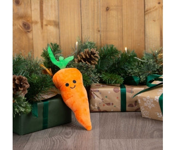 Ancol Mini Katie Carrot dog toy, a small orange plush carrot with green leaves and a smiling face, standing beside Christmas presents.