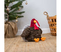 Ancol Tina Turkey plush dog toy with a shaggy brown body, red head, orange beak and large orange feet, seated beside a Christmas tree.