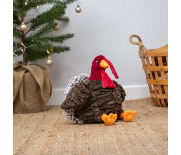 Ancol Tina Turkey plush dog toy with a shaggy brown body, red head, orange beak and large orange feet, seated beside a Christmas tree.