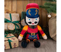 Ancol Giant Nicholas Nutcracker plush dog toy in a red uniform, tall red-and-navy hat and black boots, seated beside Christmas presents.