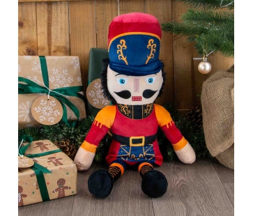 Ancol Giant Nicholas Nutcracker plush dog toy in a red uniform, tall red-and-navy hat and black boots, seated beside Christmas presents.