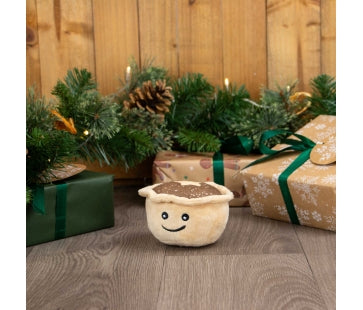 Ancol Mini Matty Mince Pie dog toy with a small cream-coloured pie body, brown top and smiling face, standing in front of Christmas presents.