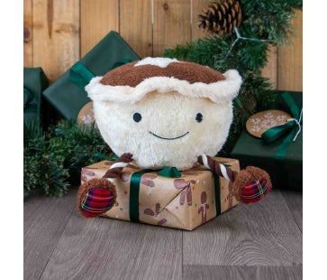 Ancol Mikey Mince Pie plush dog toy with a cream fluffy pie body, brown lid, smiling face and long legs with tartan feet, sitting on a wrapped Christmas present.
